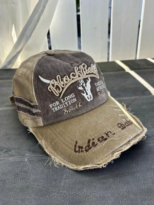 Black rebel distressed Brun och beige keps med brodyr - Säljer en brun och beige keps med broderade texter och motiv på framsidan, inklusive en tjurskalle och texten 'Blackfield', 'South', 'Indian Dakota'. Kepsen har slitna detaljer och justerbar rem bak. Materialet är bomullstyg.
