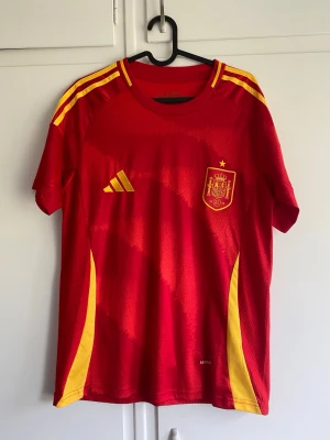 Spaniens fotbollströja röd/gul - Säljer en röd fotbollströja från Spaniens landslag med gula detaljer och emblem på bröstet. Tröjan har korta ärmar, rund hals och gula ränder på axlarna. Tillverkad i ett lätt och ventilerande funktionsmaterial, perfekt för träning eller match.