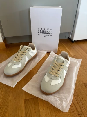 Maison Margiela sneakers beige/vit - Snygga sneakers från Maison Martin Margiela i beige mocka och vitt läder med klassisk gummisula. Låga med beigea skosnören och stilren design som passar till det mesta. Perfekta för dig som gillar minimalistisk och trendig stil.