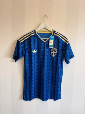 Blå Sverige fotbollströja Adidas - Säljer en blå fotbollströja från Adidas med Sveriges landslagsemblem på bröstet och gula detaljer på axlarna. Tröjan har ett mönster i olika blå nyanser, korta ärmar och V-ringning. Perfekt för dig som vill stötta Sverige på match eller träning.