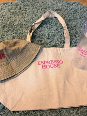 Beige tygkasse Espresso House - Säljer en beige shoppingväska i tyg från Espresso House med rosa logga tryckt på framsidan. Väskan har två långa handtag och är perfekt för att bära med sig böcker, matlåda eller annat du behöver under dagen. Plus stor mugg o solhatt