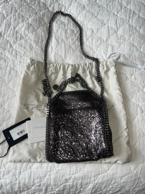 Unik Stella McCartney väska - Unik Stella McCartney Falabella bag, liten. Väskan är mörkt silvrig och glittrig med en cross body kedja och två handtag så man kan hålla i väskan också. Etikett och dustbag medföljer, knappt använd. Pris kan diskuteras!
