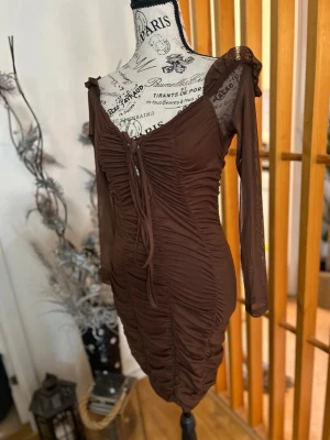 Brun meshklänning med snörning - Snygg brun kortklänning i mesh med långa ärmar och v-ringning. Klänningen har rynkade detaljer längs hela framsidan och snörning vid urringningen för extra stil. Perfekt för dig som gillar trendiga och figurnära plagg.