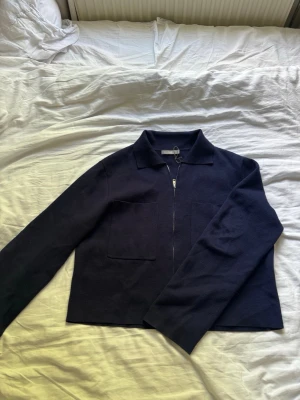 Mörkblå stickad jacka från Åhléns - Snygg mörkblå stickad jacka från Åhléns med krage och dragkedja framtill. Jackan har två stora fickor på framsidan och en rak, enkel passform som passar till många olika stilar. Finns en liten defekt. Två små gröna fläckar av något slag. Vet ej om det går bort i tvätt men ingen som syns mycket!! 