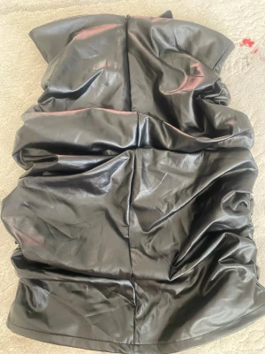 Svart kort klänning skinimitatiom - Säljer en svart  kort kjol i syntetmaterial med coolt rynkad design. Kjolen har en tight passform och ger en edgy look, perfekt för dig som vill sticka ut. Passar till dig som gillar trendiga och unika plagg.
