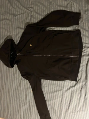 Svart hoodie med dragkedja och brodyr - Säljer en svart hoodie med dragkedja och huva. Tröjan har en liten gul broderad logga på bröstet och två fickor framtill. Tillverkad i mjukt material som känns skönt mot huden. Perfekt för en chill och avslappnad stil.