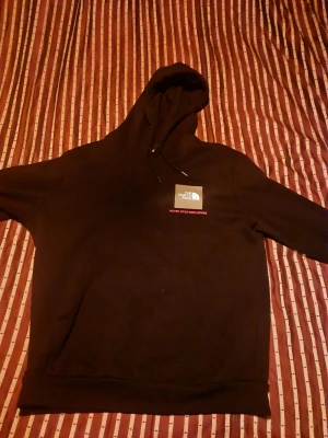 Svart hoodie från The North Face - Svart hoodie från The North Face med huva och magficka. På bröstet finns en vit logga och texten 'Never Stop Exploring' i rött. På ärmen syns en rosa logga. Perfekt för en avslappnad och cool stil.