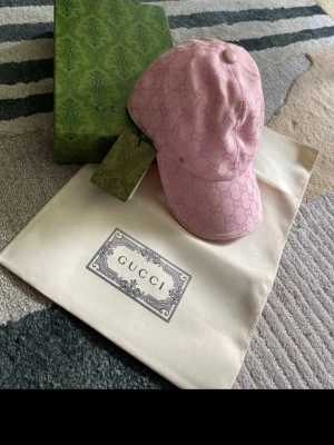 Rosa keps från Gucci med påse och låda - Säljer en snygg rosa keps från Gucci med det klassiska GG-monogrammet över hela kepsen. Kepsen har böjd skärm och justerbar rem bak. Materialet känns lyxigt och kepsen kommer med originalpåse och box.