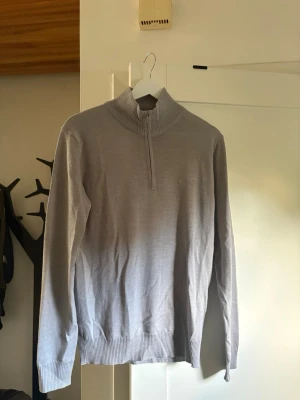 Ljusgrå halvzip från J.Lindeberg - Snygg ljusgrå stickad tröja från J.Lindeberg med halv dragkedja och hög krage. Tröjan har långa ärmar och ribbade muddar vid ärmslut och nederkant. Perfekt för lager på lager och stilrena outfits. Använt typ 5 gånger max