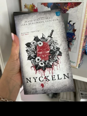 Nyckeln av Strandberg & Elfgren - Säljer boken 'Nyckeln' av Mats Strandberg och Sara Bergmark Elfgren. Omslaget är grått med en röd dödskalle, blommor och ett svärd, vilket ger en mörk och mystisk känsla. Perfekt för dig som gillar fantasy och spänning.