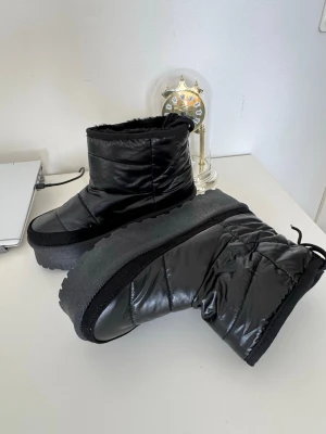 Svarta moon boots med platåsula - Säljer ett par svarta moon boots med hög platåsula och quiltad design. Bootsens utsida är i glansigt syntetmaterial och insidan är fodrad för extra värme. De har dragsko upptill för bättre passform och en chunky look som sticker ut.