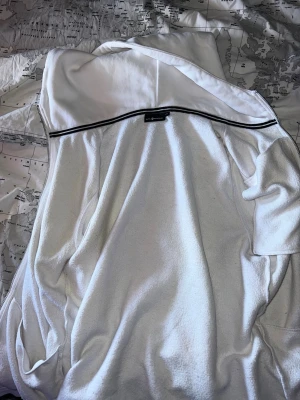 Vit hoodie med dragkedja och huva - Säljer en vit hoodie med dragkedja framtill och huva. Hoodien har en bröstficka med dragkedja och tryckt text på bröstet. Tillverkad i mjukt material som känns skönt mot huden. Perfekt för en avslappnad och sportig stil.
