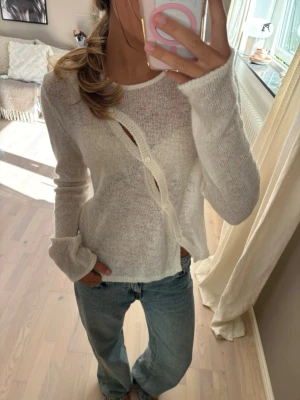 Vit asymmetrisk stickad tröja - Säljer en tunnstickad, vit tröja med lång ärm och unik asymmetrisk knäppning framtill. Tröjan har en lätt transparent look och avslappnad passform, perfekt att styla med jeans för en trendig vardagsoutfit.