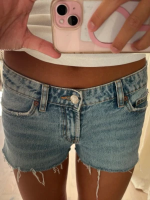 Blå jeansshorts med fransig kant - Säljer ett par ljusblå jeansshorts med rå, fransig kant och klassisk femficksmodell. Shortsen har låg midja, bälteshällor och knappgylf. Perfekta för sommaren och ger en avslappnad look.