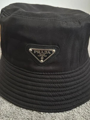 Svart bucket hat från Prada - Snygg svart bucket hat från Prada med klassisk logga framtill. Hatten är i nyttskick och aldrig andvänts pågrund av storlek. Kan gå med på att byta hatt mot hatt om de är nått du skulle ha.
