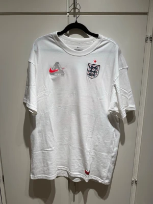 Vit England x Palace x Nike t-shirt - Vit t-shirt med England-emblem, Nike-logga och Palace-tryck framtill. Stor grafisk print på ryggen med röda lejon, Palace- och Nike-logga. Klassisk passform och rund hals. Perfekt för dig som gillar streetwear och fotbollsinspirerad stil. Kvitto/orderbekräftelse finns!