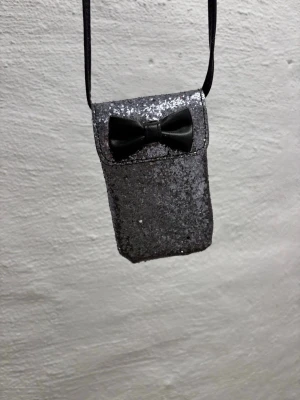 Glittrig svart mobilväska med rosett - Säljer en liten axelväska i svart med glittrig yta och en söt svart rosett på locket. Väskan har en smal axelrem och är perfekt för att bära småsaker. Materialet är troligtvis syntet med glitterdetaljer. Supersnygg accessoar för dig som gillar att sticka ut.