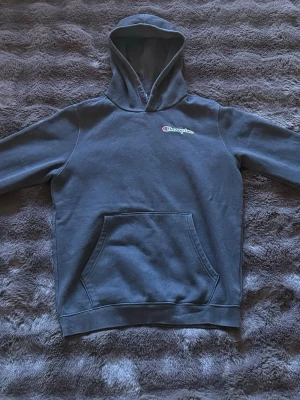 Svart hoodie från Champion - Mörkgrå hoodie från Champion med broderad logga på bröstet. Klassisk känguruficka framtill och huva. Perfekt för en chill och avslappnad stil. Mjuk bomullskänsla och ribbade muddar vid ärmslut och nederkant.