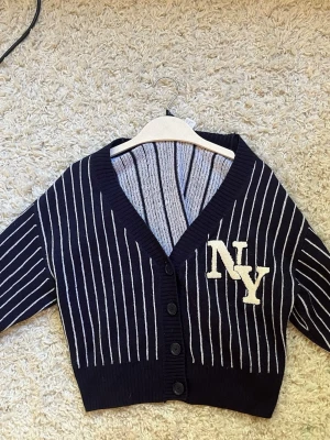 Cool mörkblå kofta med ny- tryck💗 - Snygg mörkblå cardigan med vita vertikala ränder och broderat NY-tryck på bröstet. Tröjan har v-ringning, ribbade muddar och knäppning med fem knappar framtill. Croppad modell som ger en trendig look.
