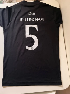 Real Madrid Bellingham matchtröja svart - Säljer en svart Real Madrid matchtröja med Bellingham och nummer 5 på ryggen. Tröjan har vita tryck med klubbmärke, sponsor och Y-3 logga framtill samt en signaturdetalj. Tillverkad i ett lätt och sportigt material, perfekt för fotboll eller träning.