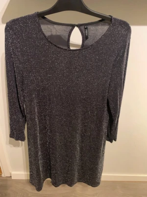 Glittrig svart klänning med 3/4-ärm - Snygg svart klänning med glittrigt tyg och 3/4-ärm. Rund halsringning och en liten öppning med knapp i nacken. Materialet är stretchigt och skimrar i silver. Perfekt för dig som vill sticka ut lite extra.
