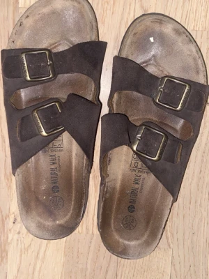 Bruna sandaler med dubbla spännen - Snygga bruna sandaler med två breda remmar i mocka och justerbara metallspännen. Fotbädden är formad för extra komfort och sulan har ett mönstrat grepp. Perfekta för en avslappnad stil under varma dagar.