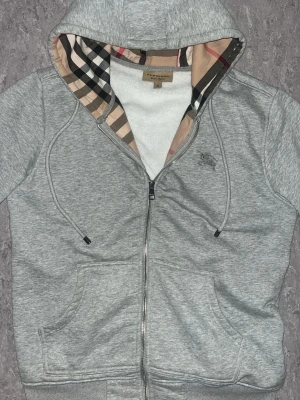 Grå hoodie med Burberry-kofta - Grå zip-hoodie med klassiskt Burberry-rutigt foder i huvan. Mjuk bomullskänsla, dragkedja framtill och två fickor. Perfekt för en stilren och avslappnad look. Huvan har det ikoniska rutmönstret i beige, svart, vitt och rött.