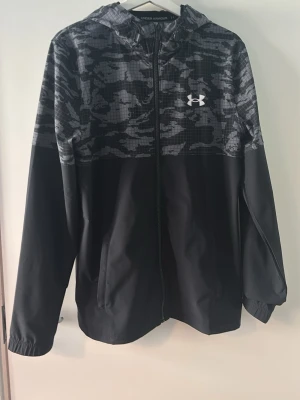 Under armour windbreaker  - köpt för 1200kr på JD. Snygg svart vindjacka från Under Armour med grått camomönster upptill och huva. Jackan har dragkedja framtill, två sidofickor och elastiska muddar vid ärmsluten. Perfekt för blåsiga dagar och sportiga outfits.