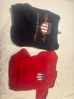 Två GANT hoodies med logga - Säljer två snygga hoodies från GANT blåa är storlek M och andra L men båda passar M en i marinblått och en i rött. Båda har huva, ribbade muddar och stor broderad GANT-logga med amerikansk sköld på bröstet. Klassisk streetwear-stil och mjukt material, perfekt för dig som gillar sportig look.