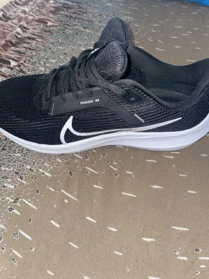 Nike Pegasus 40 svarta sneakers - Snygga svarta Nike Pegasus 40 sneakers med vit sula och vit swoosh på sidan. Ovandelen är i meshmaterial för bra andningsförmåga och skon har klassisk snörning. Perfekta för dig som gillar sportig och stilren look. (Aldrig använda, köpte fel storlek)