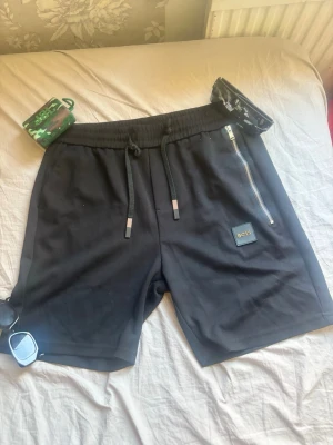 Svarta shorts från BOSS med snörning - Säljer ett par svarta shorts från BOSS med snörning i midjan och en snygg silverfärgad dragkedja på sidan. Shortsen har en stilren design med BOSS-logga på benet och är perfekta för sommaren. Sköna och lätta att matcha till olika outfits.Pis kan diskuteras.