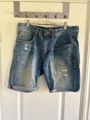 Blå denimshorts med slitningar - Säljer ett par blå denimshorts med slitna detaljer och upprullade benslut. Klassisk femficksmodell med knappgylf och casual vibe. Perfekta för varma dagar och enkel att matcha med t-shirt eller hoodie.