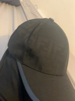 Svart keps med FF - Snygg svart keps med diskret ton-i-ton mönster över hela ytan. Klassisk böjd skärm och sexpanelers design. Materialet ser ut att vara bomull eller polyester, vilket ger en stilren och modern look.