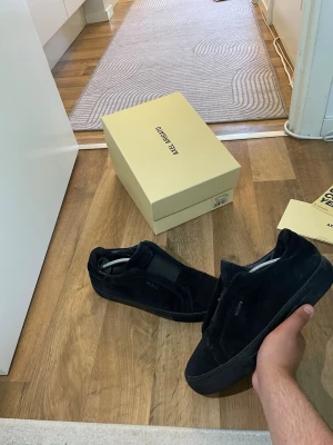 Svarta sneakers från Axel Arigato - Stilrena svarta sneakers från Axel Arigato i mocka med diskret logga på sidan. Skorna har en låg profil och svart sula för en clean look. Perfekta för dig som gillar minimalistisk design och vill ha något som funkar till det mesta.