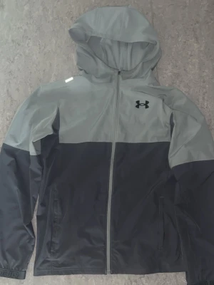 Grå vindjacka från Under Armour - Snygg tvåfärgad vindjacka från Under Armour i ljusgrått och mörkgrått. Jackan har huva, hel dragkedja framtill, två sidofickor och logga på bröstet. Tillverkad i lätt och vindtätt material, perfekt för blåsiga dagar.