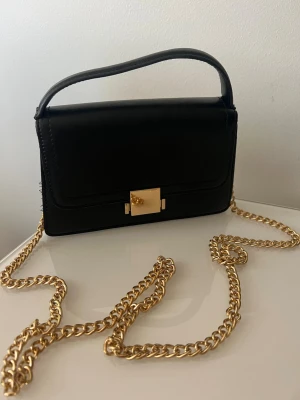 Svart handväska med guldkedja - Stilren svart handväska i slätt material med guldfärgad kedjerem och elegant guldfärgat spänne framtill. Väskan har en rektangulär form och ett kort handtag upptill, perfekt för en trendig look.