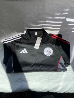 Svart Adidas träningsset med röd detalj - Säljer ett svart träningsset från Adidas med matchande tröja och byxor. Setet har vita och röda ränder på axlar och ben samt klubbmärke på både tröja och byxa. Materialet är lätt och andas, perfekt för träning eller uppvärmning.Pris kan diskuteras 