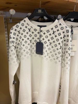 Vit Louis Vuitton sweatshirt - Snygg vit sweatshirt från Louis Vuitton med svart monogrammönster över axlar och bröst. Tröjan har rund halsringning och långa ärmar. Tillverkad i mjukt material som känns skönt mot huden. Perfekt för dig som gillar exklusiv streetwear.