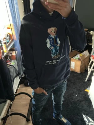 Mörkblå hoodie Polo Bear Ralph Lauren - Säljer en mörkblå hoodie från Ralph Lauren med den ikoniska Polo Bear-trycket på bröstet. Hoodien har huva och en stor magficka framtill. Perfekt för dig som gillar streetwear och vill ha något stilrent men ändå lekfullt.