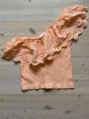 One shoulder topp - Orange one shoulder topp med volang som är köpt på zara i storlek 152cm. Tyget är stretchigt och passar de flesta. Jättefin orange eller aprikos färg.