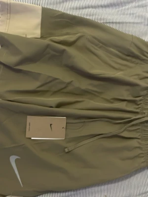 Olivgröna Nike shorts med snörning - Säljer ett par olivgröna shorts från Nike med beigea detaljer upptill. Shortsen har elastisk midja med snörning och en diskret Nike-logga framtill. Tillverkade i ett lätt syntetmaterial som passar perfekt för träning eller avslappnade dagar.