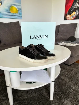 Svarta sneakers från Lanvin - Snygga svarta sneakers från Lanvin med glansig tå i lack och ovandel i mocka. Skorna har vita sulor och svarta skosnören. Klassisk låg modell som passar till det mesta och ger en stilren vibe. Det tillkommer nya lanvins snören