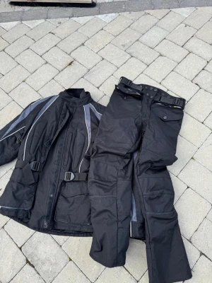 Svart motorcykelställ med reflexdetaljer - Säljer ett svart motorcykelställ med jacka och byxor. Setet har grå och vita reflexdetaljer, justerbara spännen i midjan och stora fickor på jackan. Materialet är slitstarkt och vattenavvisande.