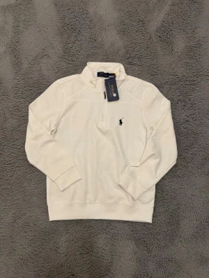 Vit halvzip tröja från Ralph Lauren - Klassisk vit tröja från Ralph Lauren med halv dragkedja och broderad marinblå logga på bröstet. Tröjan har långa ärmar, ribbade muddar och är tillverkad i mjukt material som ger en clean och stilren look.