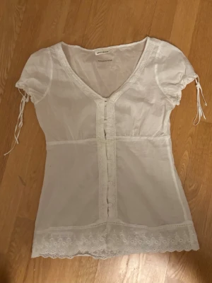 Vit blus med spetsdetaljer - Söt vit blus från DKNY Jeans med korta puffärmar och knytband. Blusen har v-ringning, knappar framtill och fina spetsdetaljer längs nederkanten och runt halsen. Tillverkad i ett tunt, luftigt material som känns romantiskt och somrigt. Blusen saknar dock en knapp näst längst upp, men de är lätt att sy på en. 