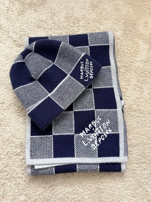 Mörkblå LV halsduk - Snygg mörkblå halsduk från LV med rutigt mönster i grått och vitt. Halsduken har broderad text och är tillverkad i mjukt material som känns lyxigt och varmt. Perfekt accessoar för att lyfta din outfit.
