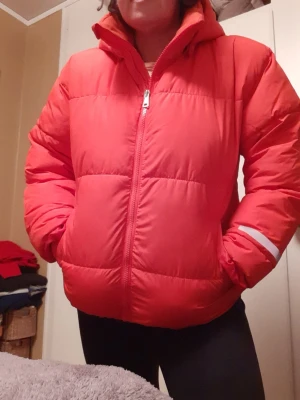Röd pufferjacka från Lindex - Cool röd pufferjacka från Lindex med hög krage och dragkedja framtill. Jackan har vattenavvisande material och en reflexdetalj på ena ärmen. Perfekt för kalla vinterdagar och håller dig varm och snygg.