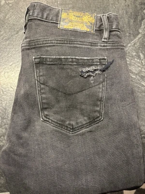 Grå slitna jeans från Crocker - Säljer ett par grå jeans från Crocker med slitningar på knäna och råa kanter vid bensluten. Jeansen har en klassisk femficksmodell och är tillverkade i ett mjukt denimtyg. Perfekta för en avslappnad och trendig look.