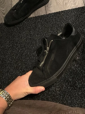 Svarta sneakers med dragkedja från Axel arigato - Stilrena svarta sneakers från Axel arigato i mocka och läder. Skorna har en unik dragkedja på ovansidan istället för klassiska snören och en svart gummisula. Använda fåtal gånger och är i mycket bra skick. Storlek 40. Pris kan diskuteras