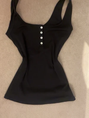 Svart ribbad linne med knappar - Säljer en svart ribbad linne med breda axelband och fyra vita knappar framtill. Toppen har en figurnära passform och är tillverkad i ett stretchigt material som sitter snyggt på kroppen. 